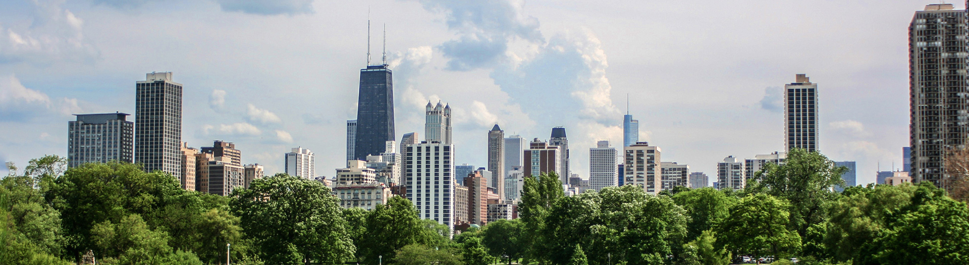Panoramic view of a Chicago skyline with a mix of tall skyscrapers and smaller buildings. Lush green trees are in the foreground.